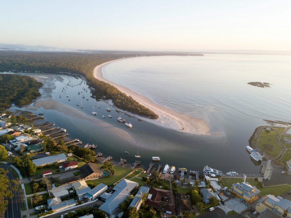 Aerial view of an inlet at Jervis Bay with moored boats, a sandbar and residential streets along the shoreline