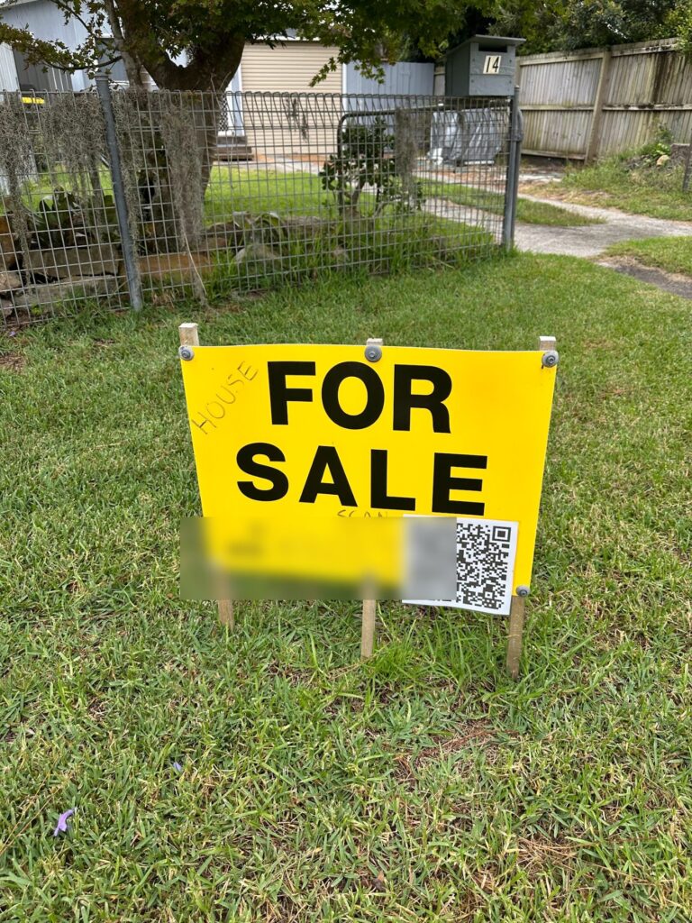 A handwritten yellow for sale sign staked on a front lawn with a QR code taped to it